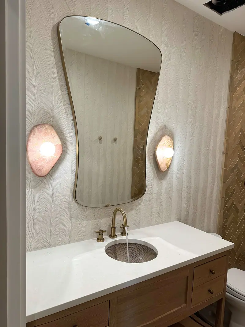Tocador de madera con encimera blanca y grifo de latón bajo un espejo curvo entre dos apliques rosados en forma de concha, sobre azulejo blanco texturizado