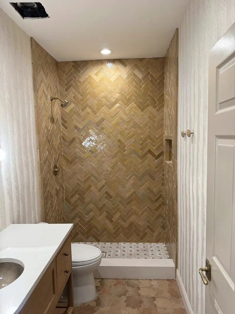 Ducha abierta con una pared de azulejo brillante en espiga verde oliva y dorado, un cabezal de ducha de latón y un piso de mosaico blanco, junto a un tocador de madera