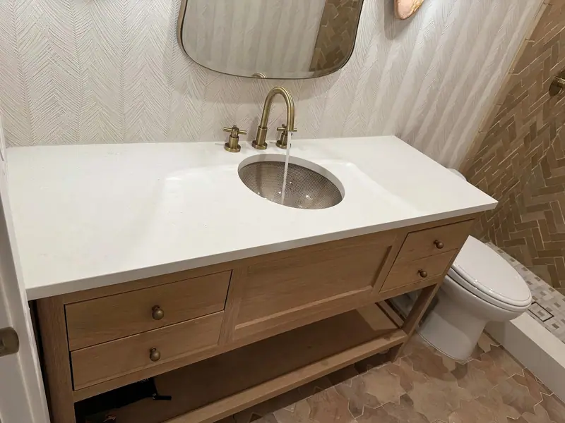 Tocador de madera clara con repisa abierta, encimera blanca, lavabo empotrado y grifo de latón, junto a la pared de la ducha en espiga