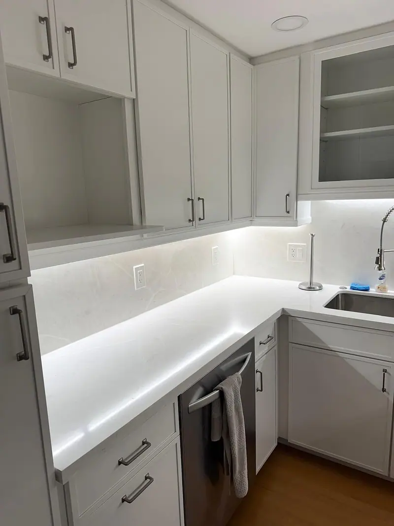 Esquina de una cocina con gabinetes gris claro estilo shaker, encimera y salpicadero blancos iluminados bajo los gabinetes, un lavavajillas de acero inoxidable y piso de madera