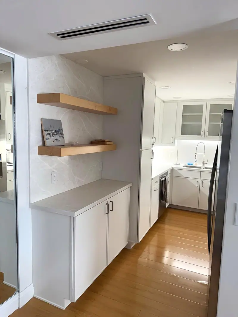 Repisas flotantes de madera en una pared blanca junto a gabinetes gris claro, con vista a una cocina tipo pasillo con piso de madera