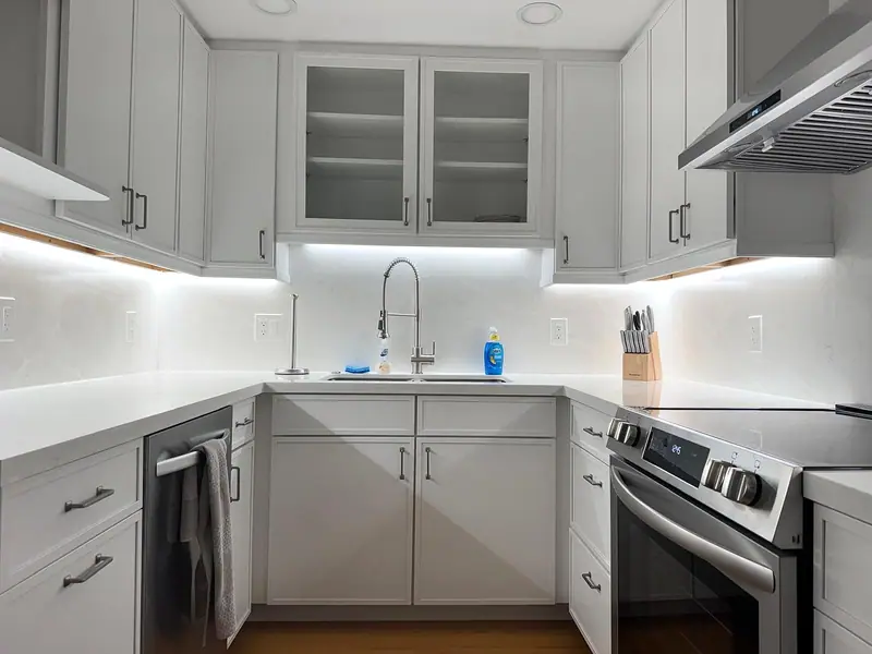 Cocina con gabinetes gris claro estilo shaker, gabinetes superiores con puertas de vidrio sobre el fregadero, encimera y salpicadero blancos iluminados bajo los gabinetes, y una estufa y campana de acero inoxidable