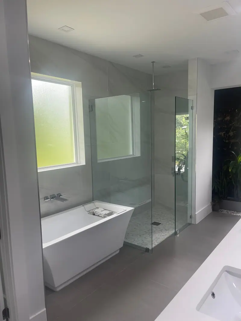 Bañera independiente rectangular con grifo de pared bajo una ventana esmerilada, junto a una ducha de vidrio sin marco con regadera de techo tipo lluvia, sobre azulejo gris grande en el piso