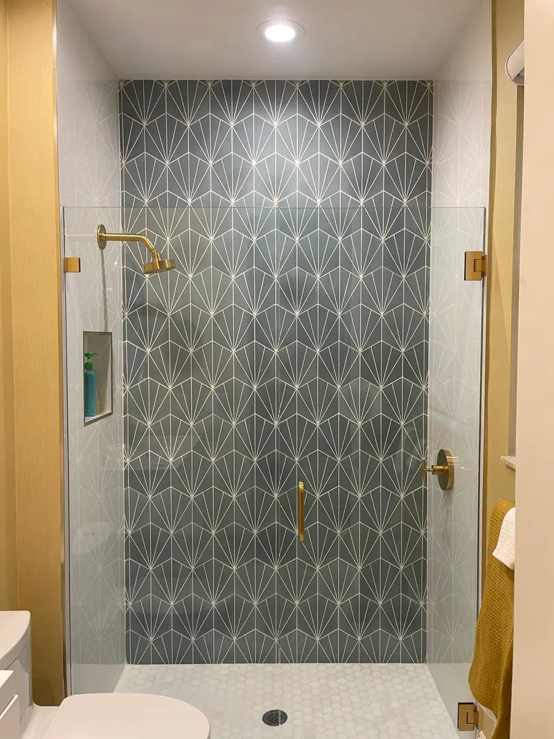 Ducha abierta con una pared de azulejo geométrico gris oscuro, un piso de mosaico hexagonal blanco, griferías doradas y una puerta de vidrio sin marco