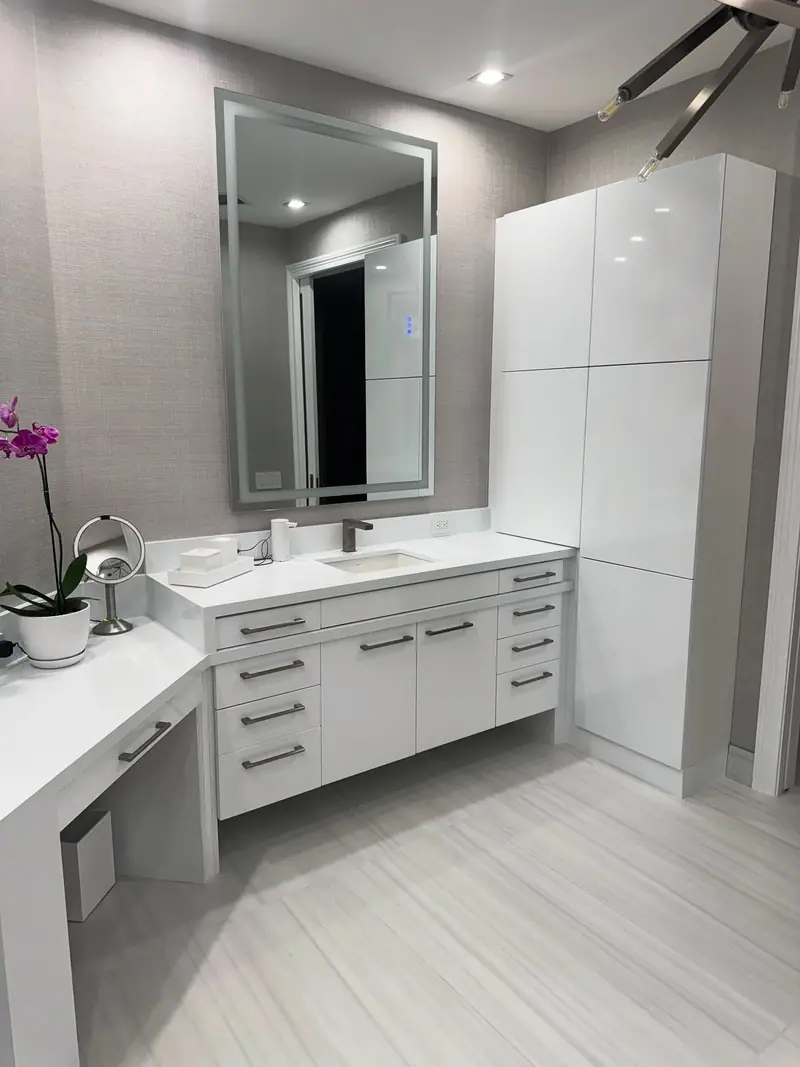 Tocador blanco brillante con espejo LED, un gabinete alto para toallas y una mesa de maquillaje, sobre papel tapiz gris texturizado