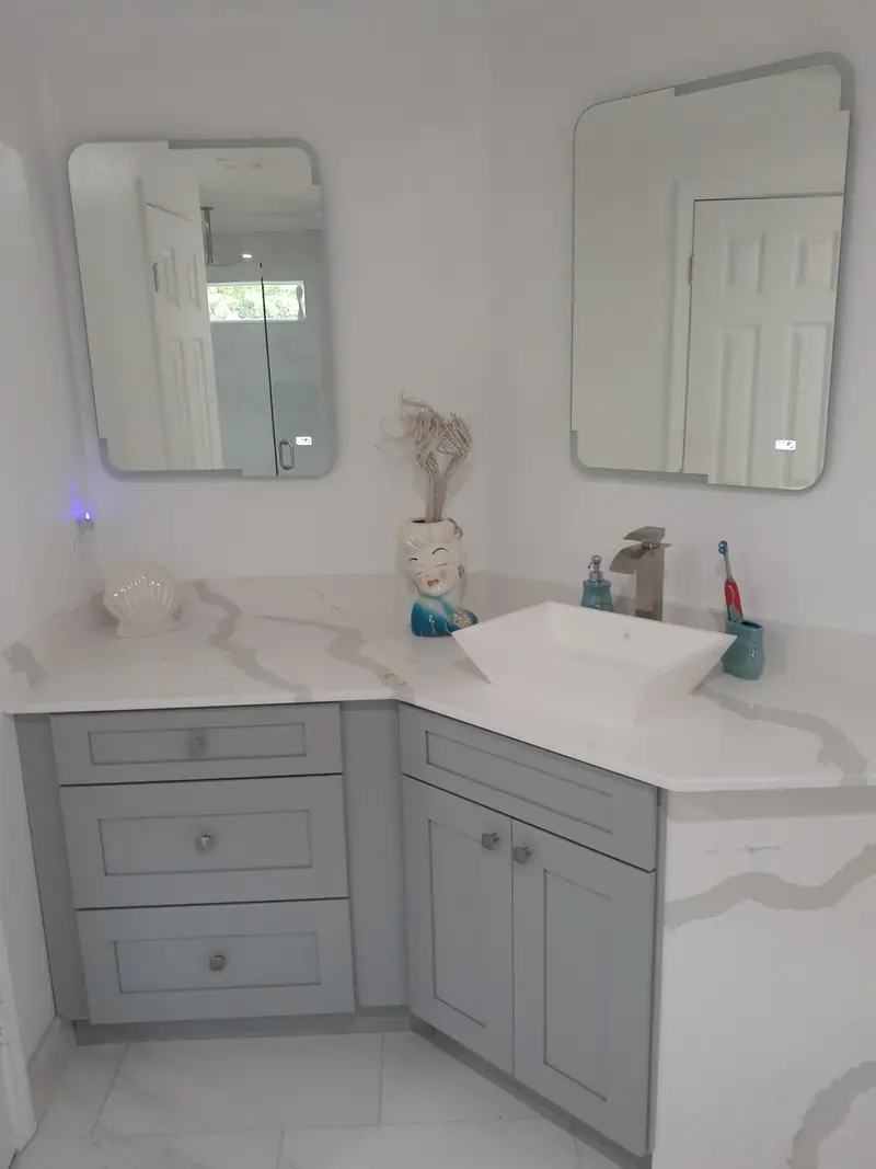 Tocador esquinero gris estilo shaker con encimera blanca veteada que baja por el costado, un lavabo blanco sobre encimera y dos espejos sin marco