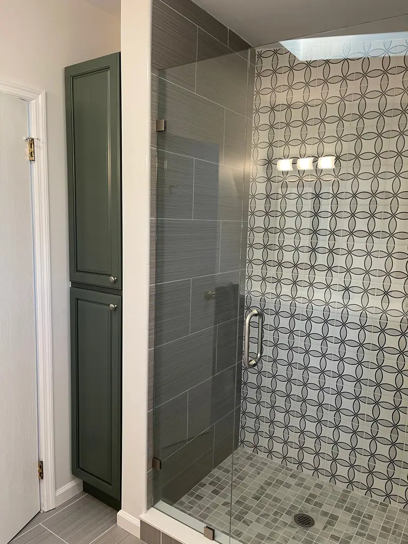 Ducha abierta de vidrio con una pared de azulejo con dibujo de círculos, azulejo gris tipo tablón y un piso de mosaico pequeño, junto a un gabinete alto verde para toallas