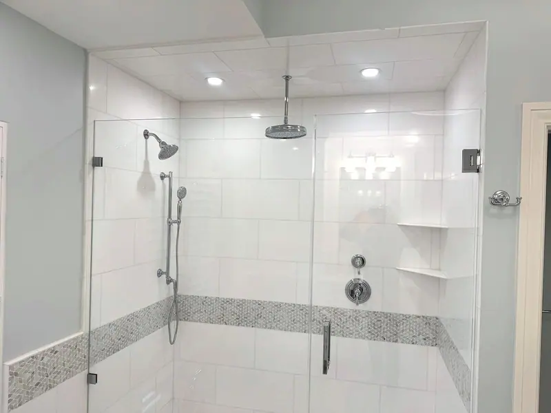 Ducha de vidrio sin marco con paredes de azulejo blanco, una franja de mosaico gris, una regadera de techo, una regadera de pared y una ducha de mano en barra deslizable