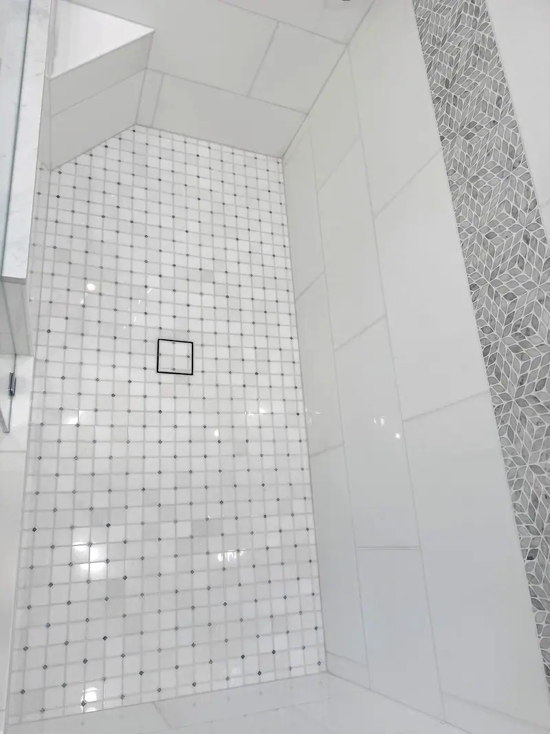 Shower floor of white mosaic with small gray dots and a square drain, with white wall tile and a gray mosaic stripe