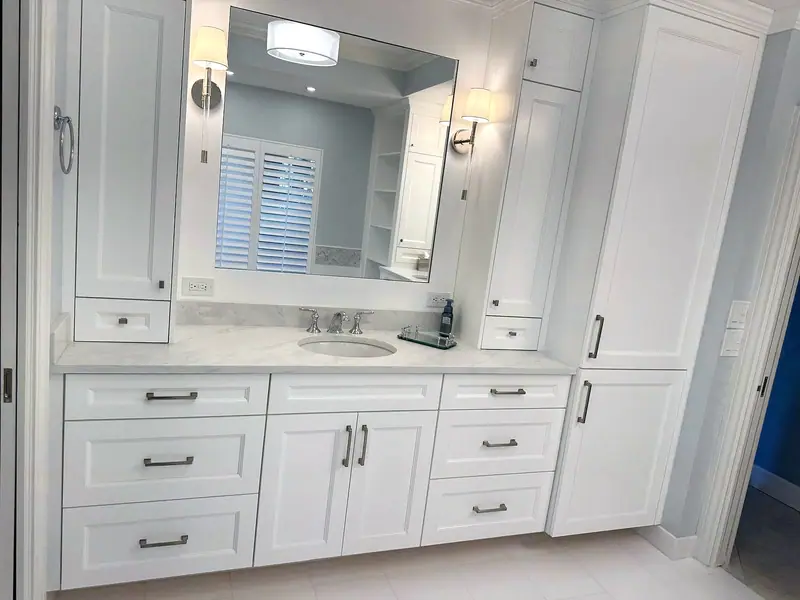 Tocador blanco estilo shaker con encimera blanca veteada, un espejo ancho entre dos apliques de pared y gabinetes altos blancos a ambos lados