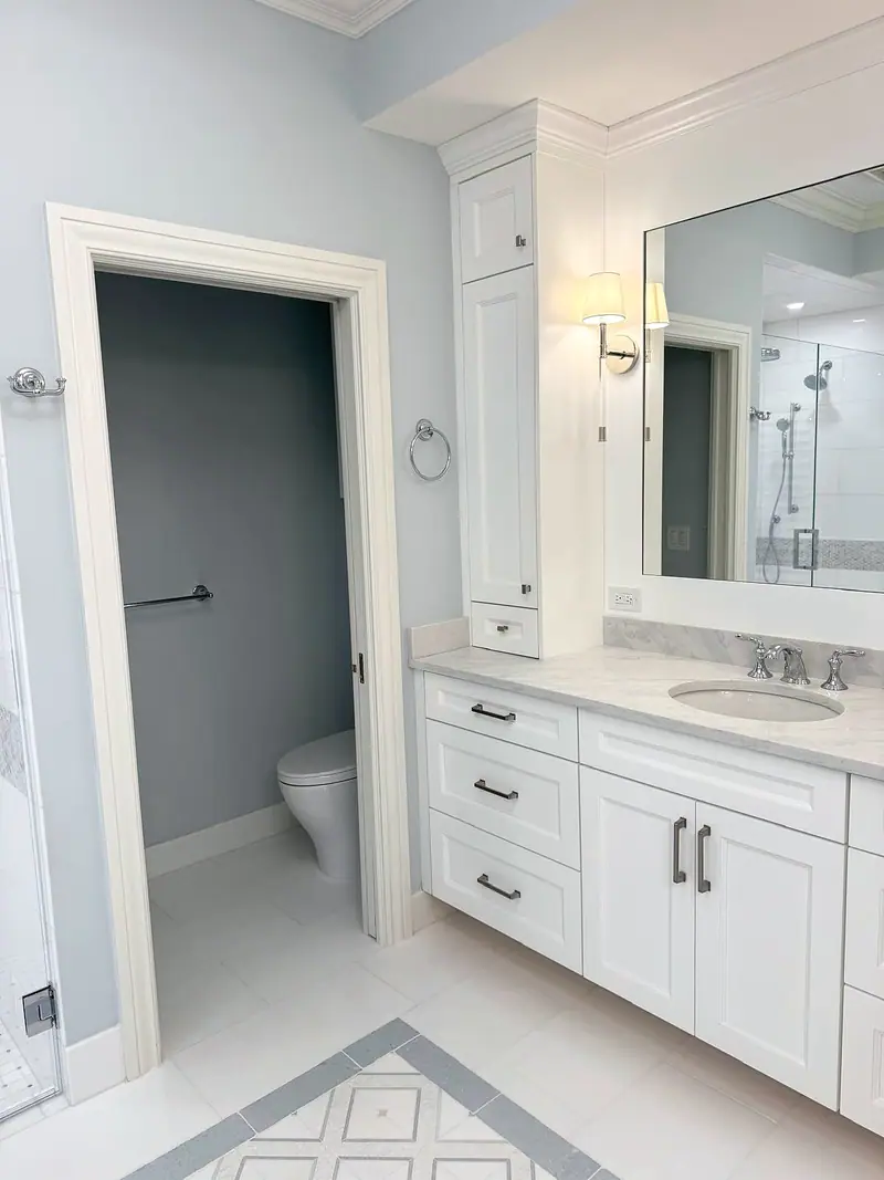 White shaker vanity with a mirror and a wall sconce, next to the door of a separate toilet room with blue-gray walls
