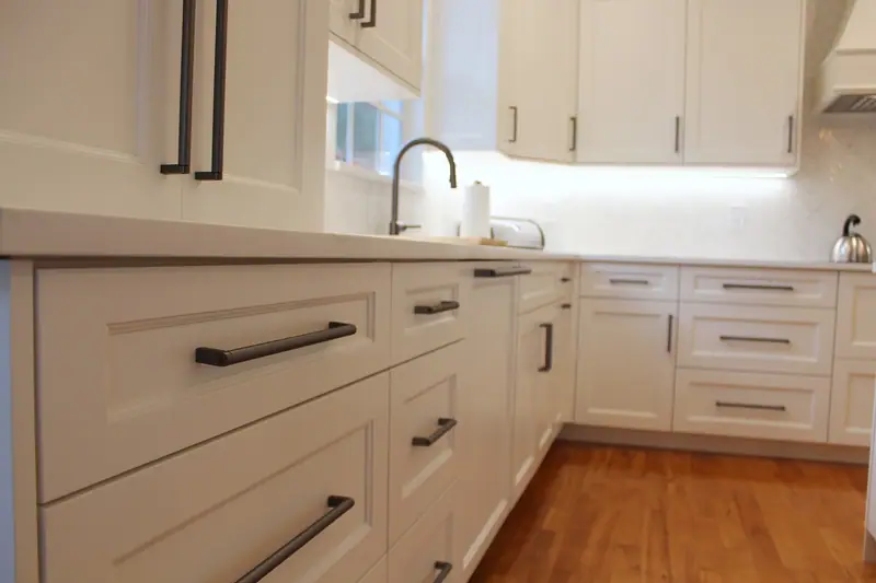 Close-up of white shaker drawers with dark bar pulls along a white quartz counter, with the sink and the backsplash beyond