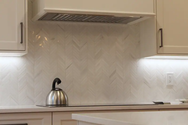 White marble chevron mosaic backsplash with soft gray veins under a range hood, lit by strips under the cabinets, with a kettle on the cooktop