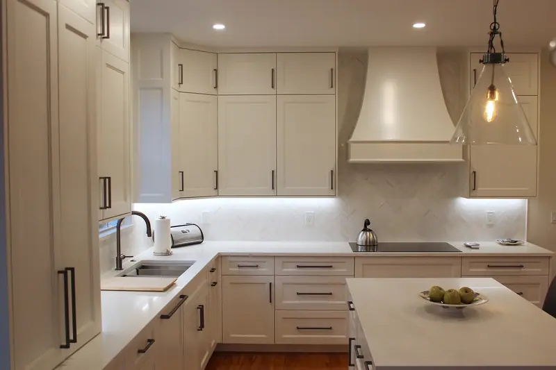 Curved range hood over a black cooktop, between white shaker cabinets, with lighting under the cabinets and a white marble chevron mosaic backsplash