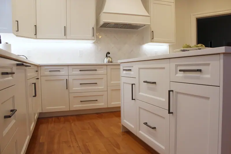 Corner of white shaker base cabinets with drawers and dark bar pulls on a wood floor, with the range hood in the background