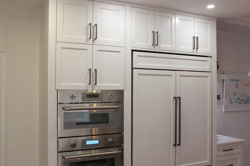 Refrigerator behind white cabinet panels with long dark pulls, beside two stacked stainless wall ovens, under white shaker cabinets
