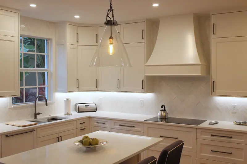 Sink with a dark pull-down faucet under the window, white shaker cabinets on both sides, a curved range hood and a glass pendant with an exposed bulb