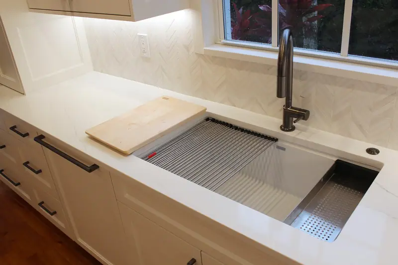 Gran fregadero tipo estación de trabajo de acero inoxidable en una encimera de cuarzo blanco, con una tabla de cortar de madera, una rejilla enrollable y un colador, bajo una grifería extraíble oscura junto a la ventana
