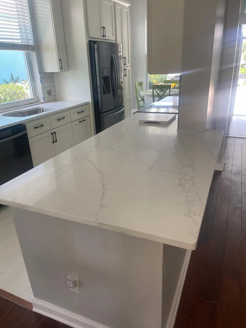 Encimera blanca veteada en una península de cocina, con gabinetes blancos estilo shaker, herrajes negros, refrigerador y lavavajillas negros, una ventana sobre el fregadero y piso de madera oscura