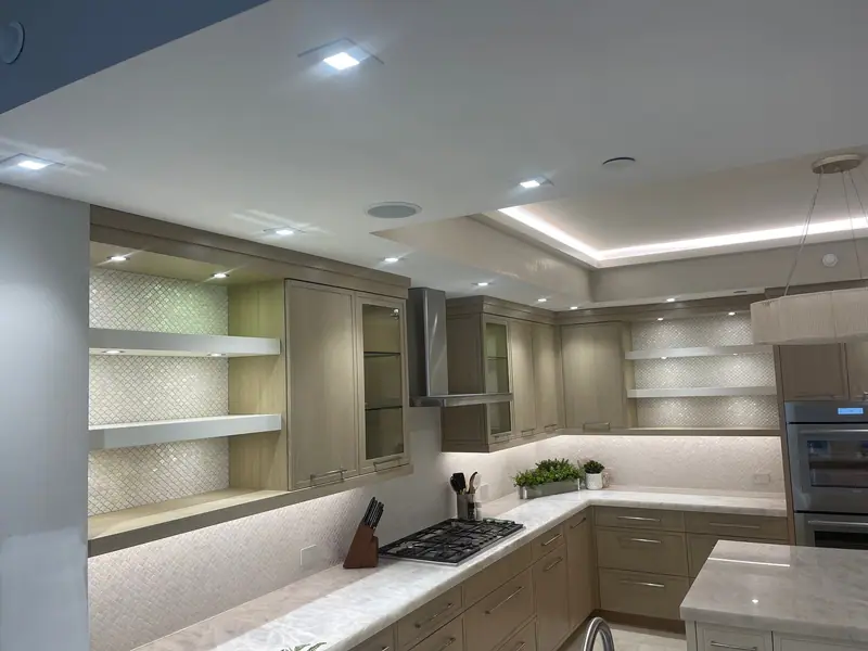 Esquina de cocina con repisas flotantes blancas sobre una pared de mosaico blanco perlado, gabinetes de madera clara con puertas de vidrio, una campana de acero inoxidable sobre una estufa de gas empotrada e iluminación bajo los gabinetes