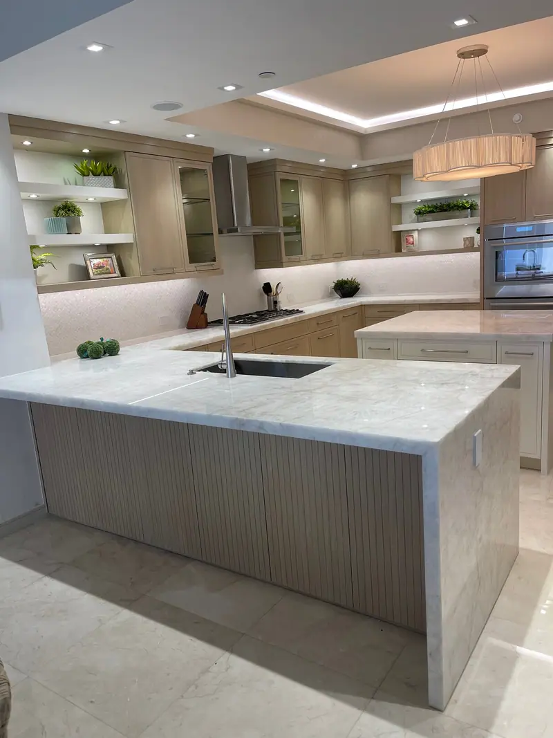Cocina con gabinetes de madera clara, una península de piedra en cascada con base acanalada, repisas flotantes y una lámpara colgante de tambor bajo un techo escalonado iluminado