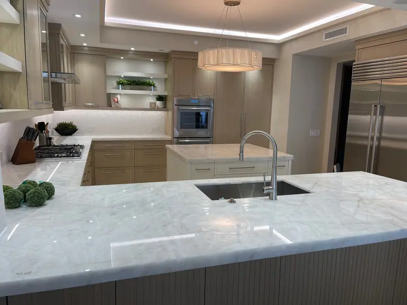 Península de piedra blanca veteada con fregadero empotrado, con vista a una isla blanca, hornos de pared, un refrigerador empotrado de acero inoxidable y una lámpara colgante de tambor bajo un techo escalonado iluminado