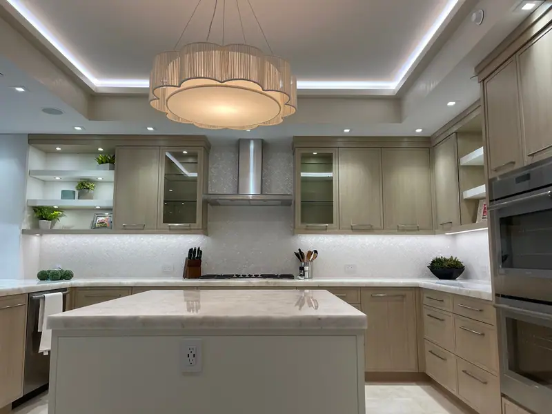 Cocina con gabinetes de madera clara, una isla blanca con encimera de piedra, un salpicadero de mosaico blanco perlado y una lámpara colgante de tambor bajo un techo escalonado iluminado