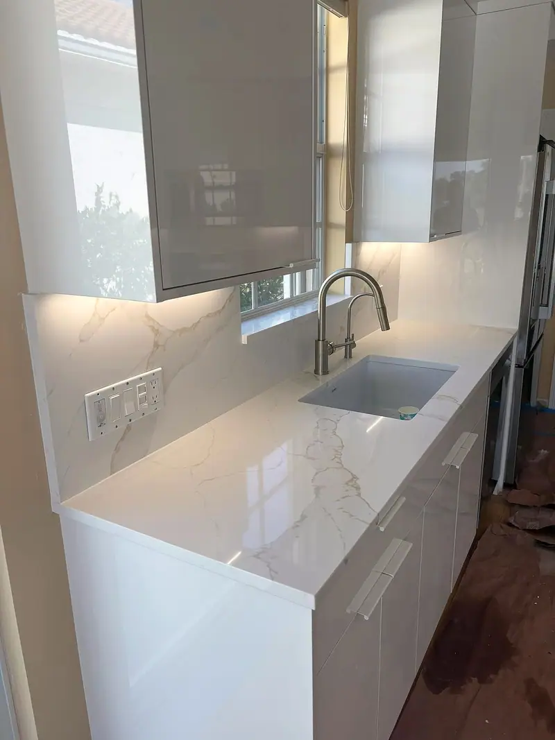 Esquina de cocina con gabinetes blancos brillantes, una encimera blanca con vetas grises que sube por el salpicadero y un fregadero empotrado bajo una ventana