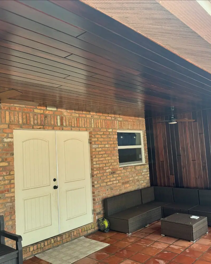 Patio techado con un techo de tablas de madera color marrón rojizo, paredes de ladrillo, puertas dobles blancas, una ventana y sofás de exterior sobre un piso de baldosas rojas