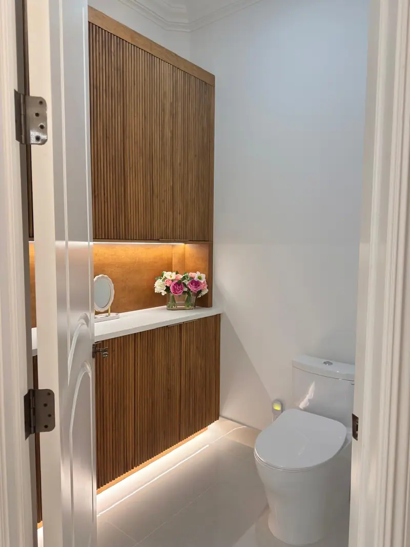 Cuarto de inodoro con gabinetes de madera acanalada de piso a techo, un nicho iluminado y una encimera blanca