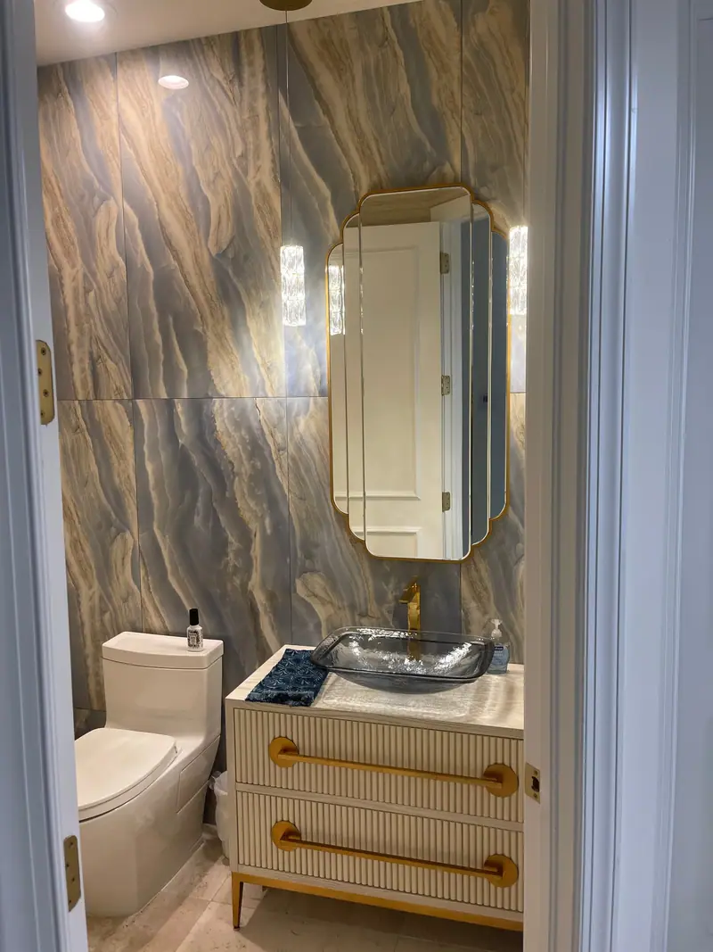 Medio baño con una pared de grandes paneles veteados en gris azulado y dorado, un espejo dorado festoneado entre colgantes de cristal, un lavabo de vidrio sobre encimera y un tocador acanalado con tiradores dorados