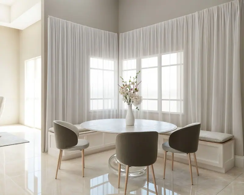 Design rendering of a breakfast nook with a round table, gray chairs and a built-in bench, by corner windows with sheer curtains