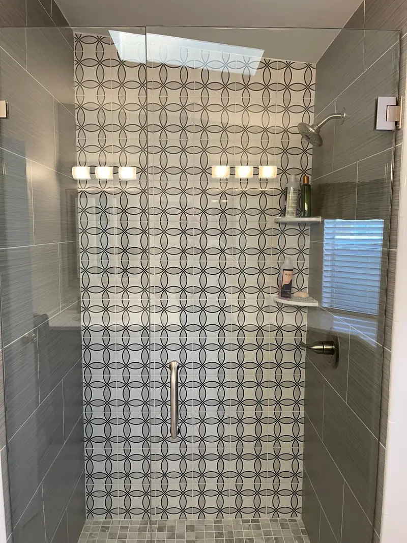 La misma ducha de azulejo con círculos vista de lado, con repisas en la esquina, una ventana y una regadera tipo lluvia tras el vidrio sin marco, bajo un tragaluz