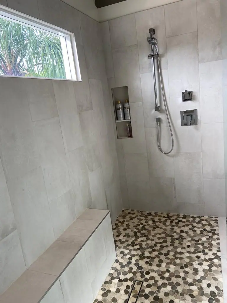 Ducha abierta con azulejo grande gris claro, un piso de piedras con desagüe lineal, un banco empotrado, un nicho en la pared, una regadera de mano en barra deslizante y una ventana arriba