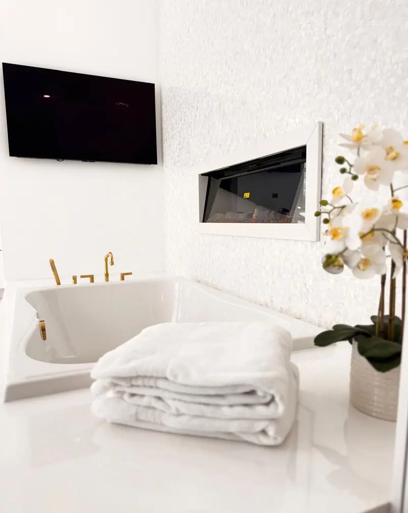 Bañera de remojo empotrada con griferías doradas junto a una chimenea lineal en una pared de mosaico blanco perla, con un televisor arriba
