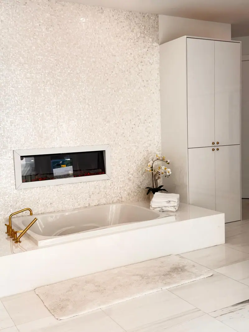 Bañera empotrada sobre una plataforma blanca elevada con griferías doradas, contra una pared de mosaico blanco perla con una chimenea lineal, junto a un gabinete alto blanco brillante con perillas doradas