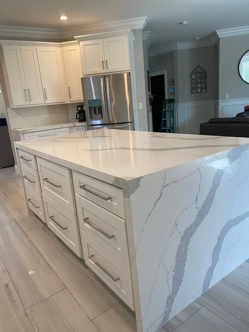 Isla de cocina blanca estilo shaker con encimera en cascada de piedra blanca con vetas grises marcadas, frente a un refrigerador de acero inoxidable