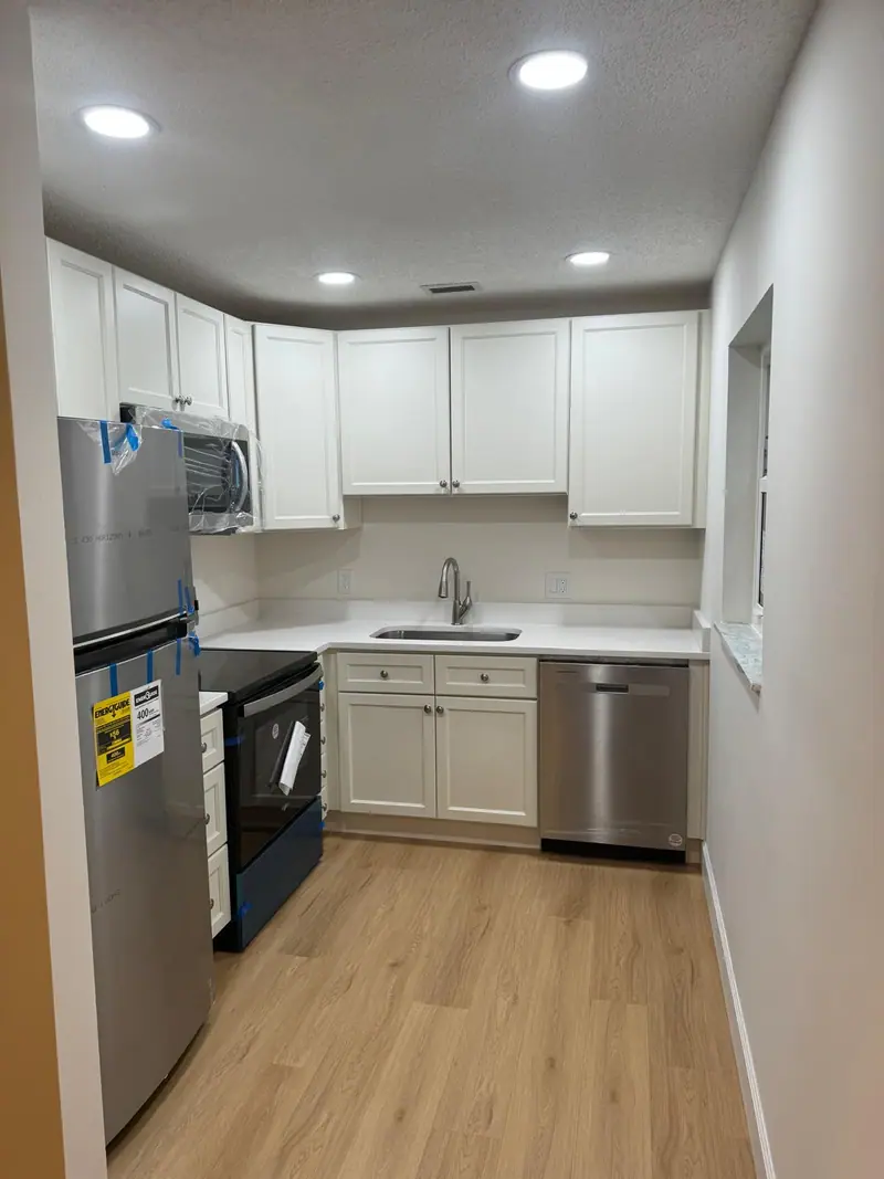 Cocina con gabinetes blancos estilo shaker, refrigerador, microondas y lavavajillas de acero inoxidable, una estufa negra, luces empotradas y piso tipo madera claro